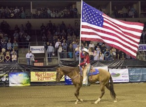 The rodeo queen is riding a brown horse and carrying an American flag.